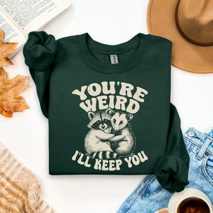May include: Dark green sweatshirt with the text "YOU'RE WEIRD I'LL KEEP YOU" in white, featuring a raccoon and opossum hugging. The sweatshirt is folded, with a hat, jeans, and a book in the background.