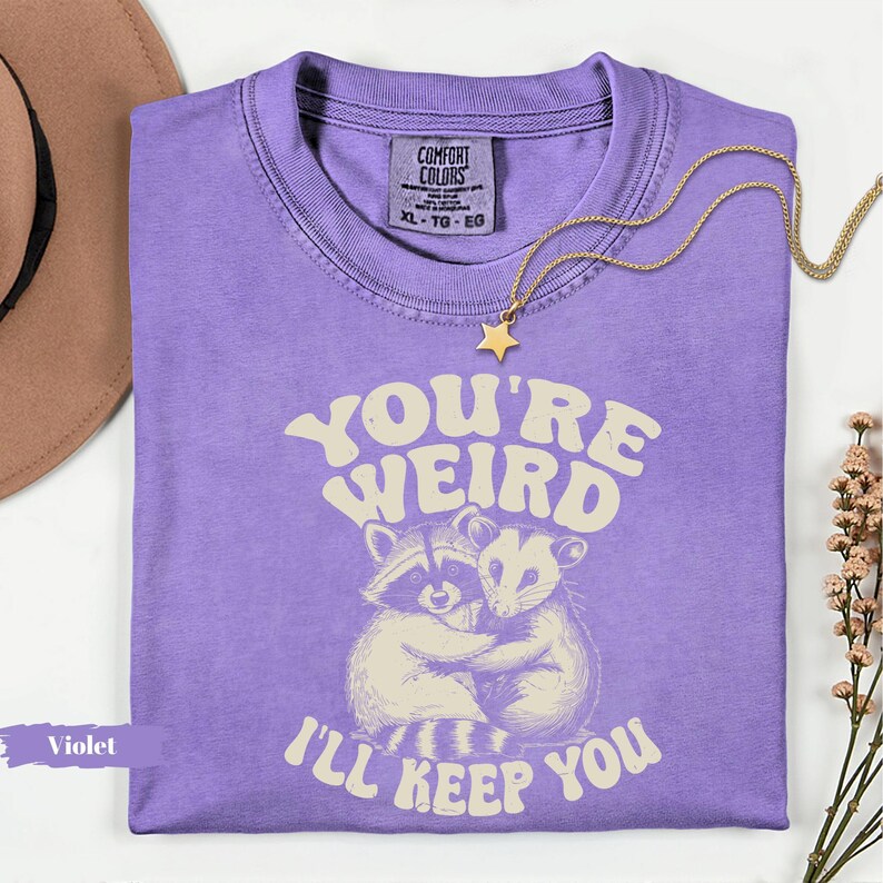 May include: Violet-colored t-shirt with the text "YOU'RE WEIRD I'LL KEEP YOU" and a graphic of two hugging raccoons. A gold star pendant necklace and a brown hat are also visible.