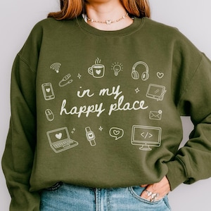 May include: Military green sweatshirt with the phrase "in my happy place" and various white icons. The icons include a laptop, coffee cup, headphones, and a computer monitor. The sweatshirt is a relaxed fit.