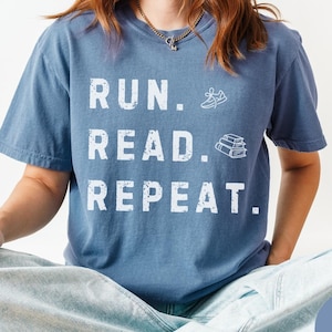 May include: A blue jean colored short-sleeve t-shirt featuring white distressed text that reads RUN. READ. REPEAT. The design includes a small white graphic of a running shoe next to the word RUN and a stack of three books next to the word READ.