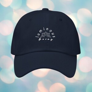 May include: A navy blue baseball cap with white embroidery that reads "luminous being" in a circular arc with a sun graphic inside the arc.