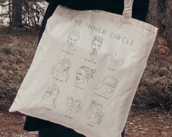 May include: A beige tote bag with black line drawings of faces and the text "THE INNER CIRCLE" printed on the front. Each face has a label underneath, such as "The Wise Lead" and "The Rebel".