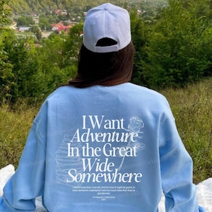 May include: A light blue sweatshirt with a white graphic print that says "I Want Adventure In The Great Wide Somewhere". The graphic also includes a rose and a stack of books. The sweatshirt is being worn by a person sitting on a blanket in a grassy area with a mountain view in the background.