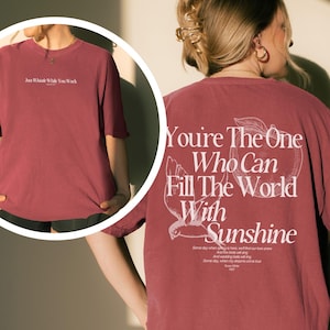 May include: A maroon oversized t-shirt with white text on the front and back. The front of the shirt reads "Just Whistle While You Work". The back of the shirt has the phrase "You're The One Who Can Fill The World With Sunshine".