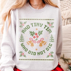 May include: A gray sweatshirt with a Christmas-themed design featuring the text "And Tiny Tim Who Did Not Die". The design is in a pixelated style, resembling a cross-stitch pattern.