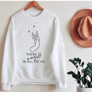 May include: White sweatshirt with a black line drawing of a hand with stars above it. The text "THERE IS magic IN ALL OF US" is printed below the hand.