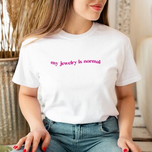 Peut inclure: Un t-shirt blanc avec le texte "my jewelry is normal" imprimé en rose.