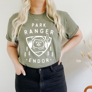 May include: A green t-shirt with a white graphic of a creature from Star Wars, a spear, and trees. The text on the shirt reads "Park Ranger Endor".