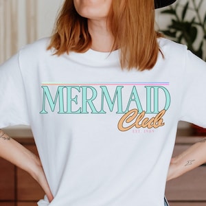Op de afbeelding: Wit T-shirt met een turquoise blauwe grafiek met de tekst "Mermaid Club Est. 1989". De grafiek heeft een regenboogstreep erboven en een perzikkleurige omtrek.