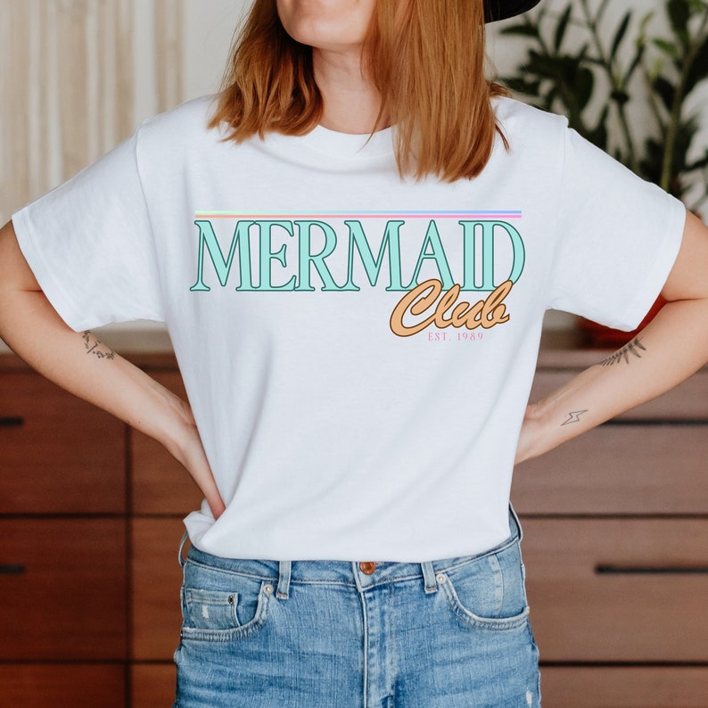 Op de afbeelding: Wit T-shirt met een turquoise blauwe grafiek met de tekst "Mermaid Club Est. 1989". De grafiek heeft een regenboogstreep erboven en een perzikkleurige omtrek.