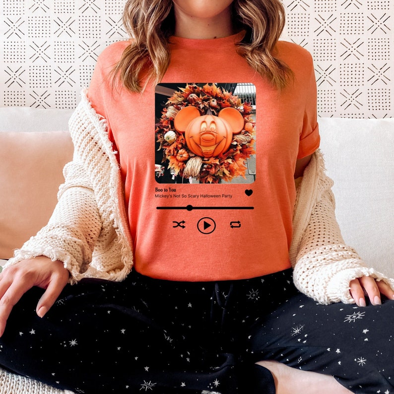 May include: Orange t-shirt with a graphic of Mickey Mouse as a pumpkin in a wreath. The graphic is in a square frame with a play button and other music controls. The text "Boo to You Mickey's Not So Scary Halloween Party" is below the graphic.