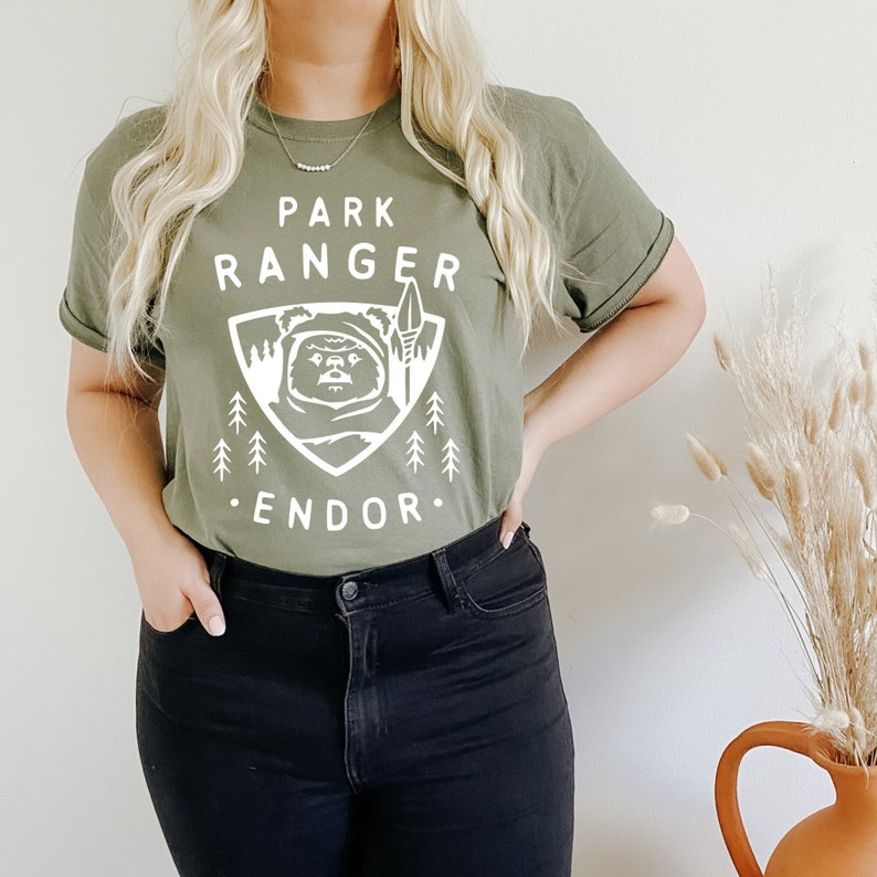 May include: A green t-shirt with a white graphic of a creature from Star Wars, a spear, and trees. The text on the shirt reads "Park Ranger Endor".