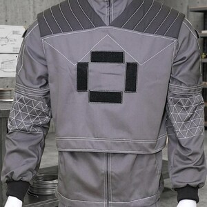 May include: Gray work uniform with a high collar, zipper, and black accents. The jacket features geometric stitching and black patches. The sleeves have black cuffs and geometric patterns. The uniform is displayed on a mannequin.