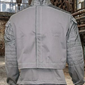May include: Gray jacket and pants set with white stitching details. The jacket has a stand-up collar, multiple pockets, and horizontal stitching on the sleeves. The pants are visible at the bottom of the image. The outfit is displayed on a mannequin.