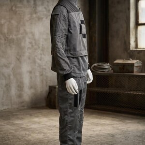 May include: A gray jumpsuit displayed on a mannequin. The outfit features a high collar, quilted shoulder details, and geometric stitching on the sleeves. Black accents are present on the chest and legs. The pants have a straight leg design. The mannequin wears white gloves and shoes.