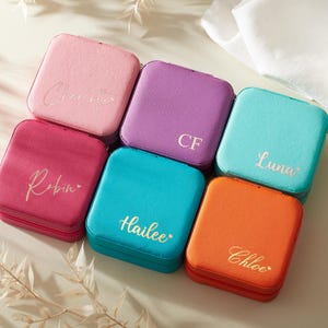 May include: A selection of personalised square jewellery cases in shades of pink, purple, teal, and orange. Each case has a name or initials in gold script. The cases are displayed on a neutral surface.