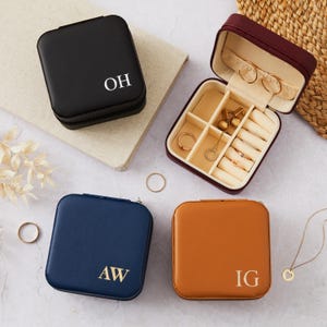 Puede incluir: Una colección de pequeñas joyeros cuadrados en negro, azul marino y marrón claro. Un joyero está abierto, revelando compartimentos y joyas. Cada joyero tiene iniciales impresas en la parte delantera. El joyero abierto es burdeos con interior beige.