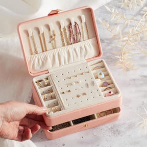 Könnte beinhalten: Geöffnete rosa Schmuckschatulle mit mehreren Fächern. Der obere Bereich enthält Halsketten, während der mittlere Bereich Ohrringschlitze hat. Die Schatulle ist mit verschiedenen Gold- und Perlenschmuck gefüllt.