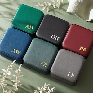 Puede incluir: Una colección de seis estuches cuadrados para joyas, de aspecto similar al cuero, en varios colores: verde, negro, burdeos, gris, azul y verde azulado. Cada estuche está grabado con iniciales doradas o blancas. Los estuches están dispuestos sobre una superficie verde.