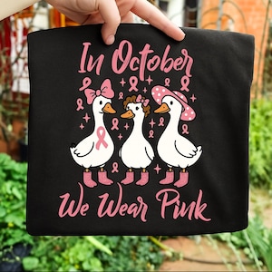 以下が含まれることがあります： ピンク色のグラフィックが特徴の黒いTシャツ。ピンク色のアクセサリーとブーツを身に着けた3羽の白いガチョウが描かれています。「In October We Wear Pink」の文字がピンク色で表示され、乳がん啓発のためのピンクのリボンも描かれています。