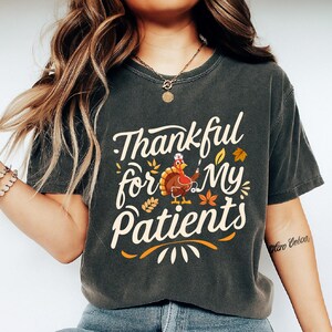 Może przedstawiać: Ciemnoszara koszulka z napisem "Thankful for My Patients" w kolorze białym i pomarańczowym. Na koszulce znajduje się rysunek kreskówkowego indyka w czepku pielęgniarki i ze strzykawką, a także jesienne liście.