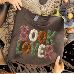 May include: A brown sweatshirt with the words "BOOK LOVER" in a knitted style. The letters are in various colors, including pink, white, orange, green, and brown. The sweatshirt is held by hands.