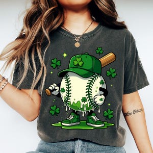 May include: Dark grey t-shirt featuring a cartoon baseball character with a green cap and shoes, holding a baseball bat. The baseball is decorated with shamrocks and green accents, with additional shamrocks scattered around the character.