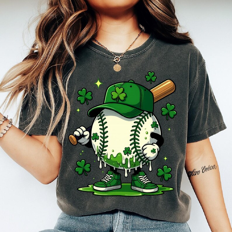May include: Dark grey t-shirt featuring a cartoon baseball character with a green cap and shoes, holding a baseball bat. The baseball is decorated with shamrocks and green accents, with additional shamrocks scattered around the character.