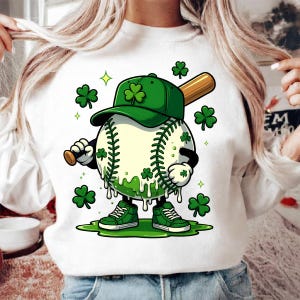 May include: White sweatshirt with a cartoon baseball character wearing a green cap with shamrocks. The baseball holds a bat and wears green trainers. Green shamrocks and dripping green accents complete the design.