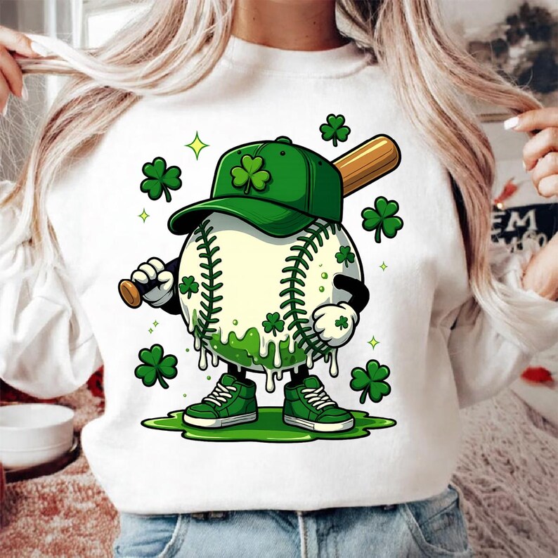 May include: White sweatshirt with a cartoon baseball character wearing a green cap with shamrocks. The baseball holds a bat and wears green trainers. Green shamrocks and dripping green accents complete the design.