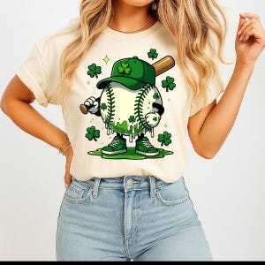 May include: Cream-coloured t-shirt featuring a cartoon baseball character with a green shamrock-themed design. The baseball wears a green cap and trainers, holding a bat. Green shamrocks and accents complete the St. Patrick's Day theme.