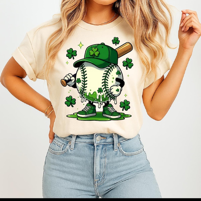May include: Cream-coloured t-shirt featuring a cartoon baseball character with a green shamrock-themed design. The baseball wears a green cap and trainers, holding a bat. Green shamrocks and accents complete the St. Patrick's Day theme.