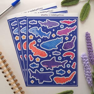 May include: Three sheets of stickers featuring various shark illustrations in shades of blue, purple, and red on a dark blue background. The stickers include text that reads "sharks + madebyalicej".
