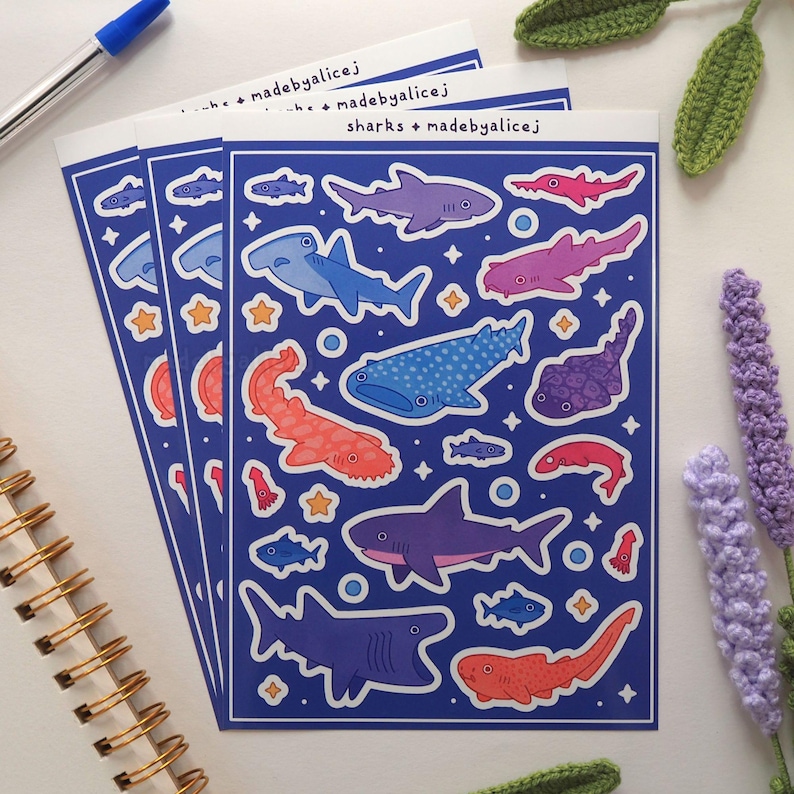 May include: Three sheets of stickers featuring various shark illustrations in shades of blue, purple, and red on a dark blue background. The stickers include text that reads "sharks + madebyalicej".