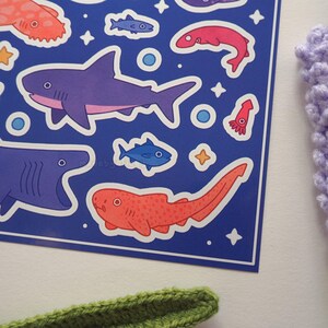 May include: A sheet of ocean-themed stickers featuring various marine life illustrations. The stickers include sharks, fish, and a squid, in shades of purple, blue, and red, set against a dark blue background. Decorative stars and circles are also present.