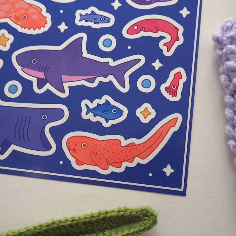 May include: A sheet of ocean-themed stickers featuring various marine life illustrations. The stickers include sharks, fish, and a squid, in shades of purple, blue, and red, set against a dark blue background. Decorative stars and circles are also present.
