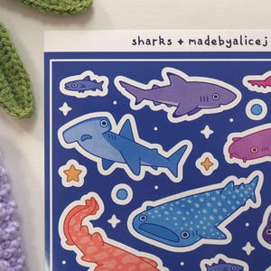 May include: A sheet of stickers featuring various shark illustrations in shades of blue, purple, and red, with white outlines. The text "sharks + madebyalicej" is printed at the top. The stickers are on a blue background.