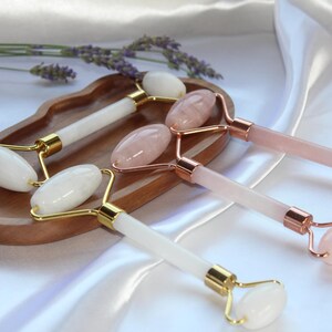 Könnte beinhalten: Mehrere Gesichts-Massage-Roller aus weißem und rosa Quarz sind auf einem Holztablett angeordnet. Die Roller haben goldfarbene und roségoldfarbene Metallakzente. Ein Zweig Lavendel liegt in der Nähe des Tabletts, vor einem Hintergrund aus weißem Satin.