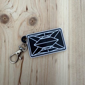 May include: A black and white geometric design keyring with a silver clasp and black rubber ring. The rectangular keyring features a black background with a white geometric pattern. It rests on a light brown wooden surface.