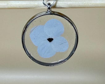 May include: A circular pendant necklace with a silver-coloured frame, showcasing a light blue flower preserved in clear resin. The flower has a dark centre and hangs from a fine silver chain.