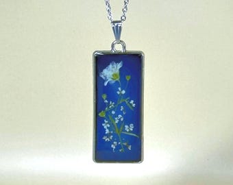 May include: A silver necklace with a rectangular pendant. The pendant displays a pressed white flower and small white blossoms against a deep blue backdrop. The pendant is encased in a silver frame.