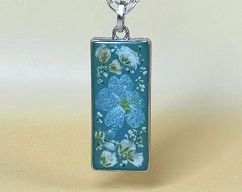 May include: A rectangular pendant with a silver-coloured frame and chain. The pendant features a teal background with a large blue flower and smaller white flowers. The flowers are encased in clear resin.