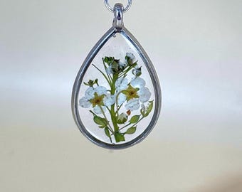 May include: A silver-framed, teardrop-shaped pendant. It contains preserved white flowers with yellow centers and green stems, set in clear resin. The pendant is suspended from a silver chain.