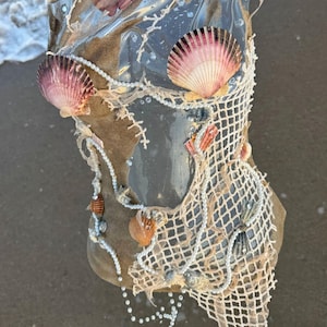 May include: A unique, handmade mermaid-inspired top. The top features a clear plastic base filled with sand, adorned with seashells, pearls, and a white net design. The seashells are pink and white.