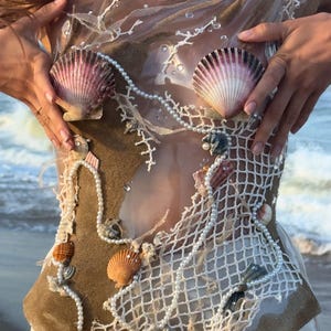 May include: A unique, handmade garment featuring a seashell design. The top is adorned with large, pink and white seashells, pearls, and a white net-like fabric. The design incorporates sand and small seashells, creating a beach-inspired aesthetic.