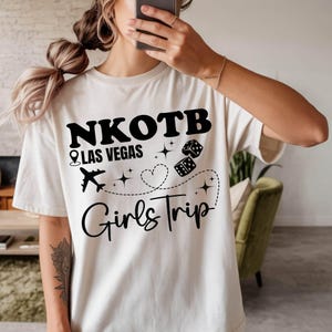 May include: Cream-colored t-shirt with the text "NKOTB Las Vegas Girls Trip" in black. The design includes a location pin, airplane, dice, and heart graphics. The shirt is a casual style.