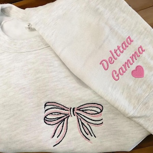 Puede incluir: Una sudadera gris claro con un diseño de lazo y el texto "Delttaa Gamma" bordado en rosa. El lazo presenta rayas negras y rosas. Un corazón rosa está bordado en la manga.