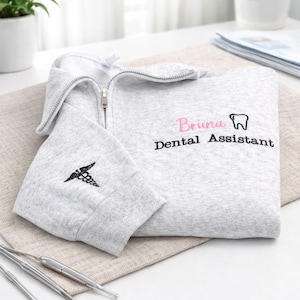Op de afbeelding: Een lichtgrijs sweatshirt met ritssluiting met de naam "Bruna" in roze en "Dental Assistant" in zwart, samen met een tandafbeelding. De linkermouw heeft een zwart caduceussymbool. Het sweatshirt is op een neutrale ondergrond gevouwen.