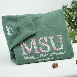 Könnte beinhalten: Grünes Sweatshirt mit gesticktem floralem "MSU"-Schriftzug, gefüllt mit rosa und weißen Blumen. Darunter steht "Michigan State University" und "Neuroscience". Der Ärmel zeigt "Alumni 2025".