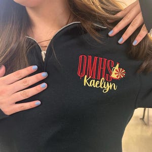 Può includere: Felpa nera con zip a un quarto di zip argentata. La felpa presenta un testo ricamato in rosso e giallo che recita "OMHS" e "Kaelyn" con una grafica da cheerleader. Un capo casual.